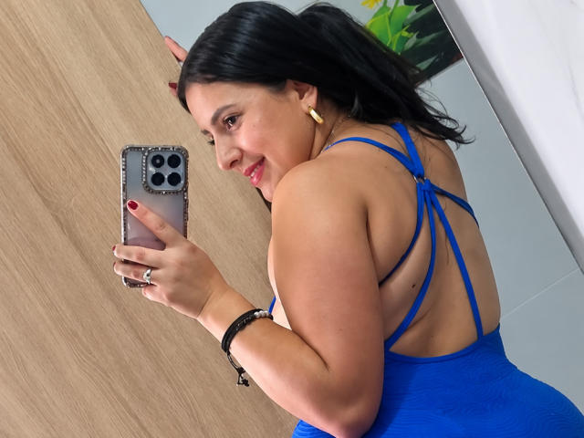AlanaDesire with black hair, red lipstick, and gold earrings, wearing a blue strappy dress, taking a mirror selfie with her phone, showing her curvy back and ass.