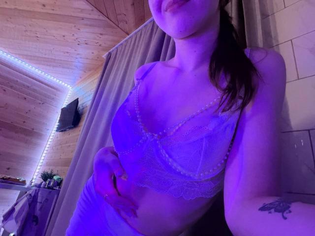 ColbyCupps in a purple-lit room, wearing a see-through lace bralette, showing her tits and a tattoo on her right arm, with her hand on her hip.