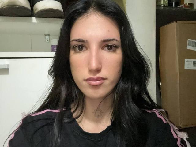 Daria Lastrape tiene el pelo largo y negro, ojos marrones y una expresión neutra, y lleva una camisa negra con rayas rosas. Está en una habitación con una caja de cartón..