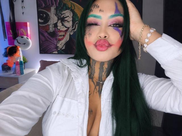LairaHearvy with green hair, white shirt, tattoos, colorful face paint, pouty pink lips, hand in hair, bracelet, and necklace, showing off her big tits.