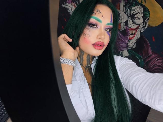 LairaHearvy with long green hair, white shirt, colorful face paint, tattoos, and green eyeshadow, holding her hair, wearing a silver bracelet, and Joker-themed background.