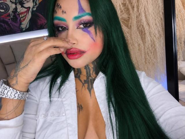 LairaHearvy with green hair, purple and blue eye makeup, red lipstick, tattoos, white shirt, ring, bracelet, touching nose, showing off her tits.