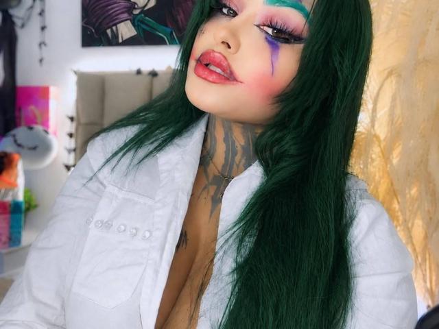 LairaHearvy mit grünen Haaren, lila und rosa Augen-Make-up, rotem Lippenstift, einem weißen Hemd, einem tätowierten Hals und freiliegendem Dekolleté. Ihr Mund ist leicht geöffnet..