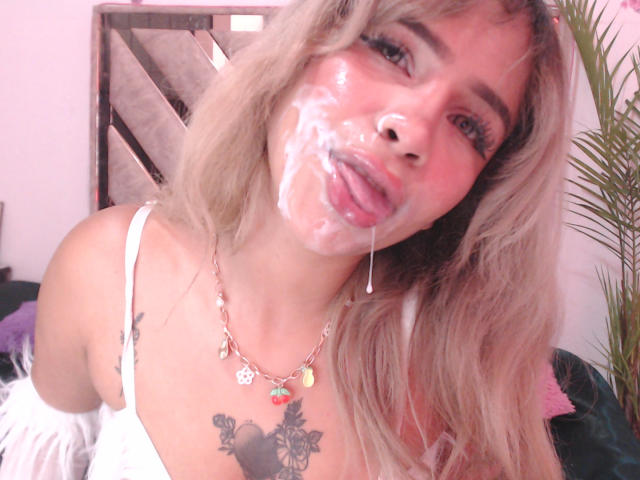 LuluDeVine com cabelo loiro, sêmen no rosto, língua para fora, babando, camiseta branca, colar, tatuagem no peito, blush rosa, olhando para a câmera, close do corpo superior..