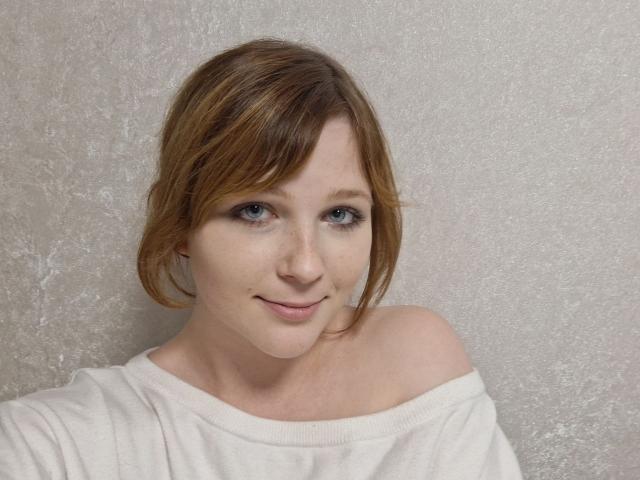 AnelleDeist with light brown hair, blue eyes, and a subtle smile, wearing a white off-shoulder shirt, has a cute, freckled face and a small nose.