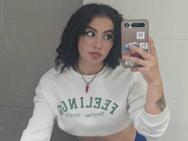 DominicTheOne with curly black hair, wearing a white crop top, holding a phone with stickers, showing a tattoo on her right arm, and a red necklace, looking in the mirror.