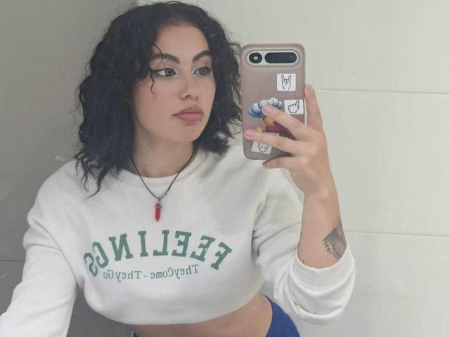 DominicTheOne with curly black hair, wearing a white "UCLA" crop sweatshirt, holding a phone with a pop socket, has a tattoo on her right arm, and a red necklace.