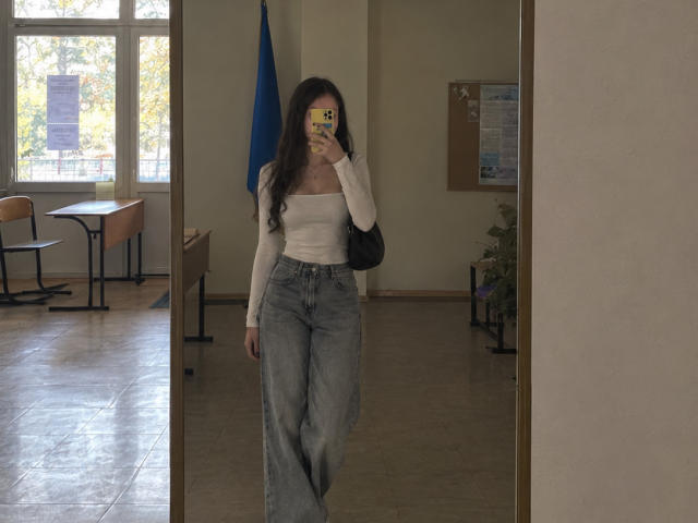 Ellaria with long brown hair, white long-sleeve top, and high-waisted blue jeans, holding phone in mirror selfie, black bag over shoulder, standing in room with windows.