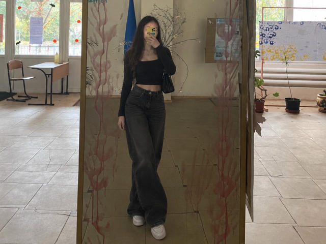 Ellaria with long brown hair, black crop top, and flared dark jeans, holding phone, standing in front of glass with red paint, wearing white sneakers, medium-ass.