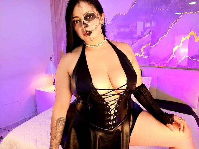 LilithAmoth with large tits, black leather dress, lace-up front, black face paint, choker, black arm sleeves, tattoo on left arm, sitting on bed, purple lighting, looking at camera.