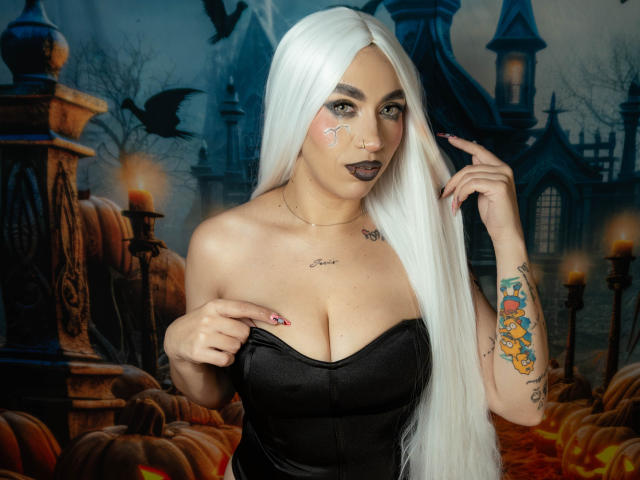 CamilleJuicyHot with long white hair, black strapless top, tattoos, face paint, dark lipstick, and painted nails, holding top, showing big tits, and looking at camera.
