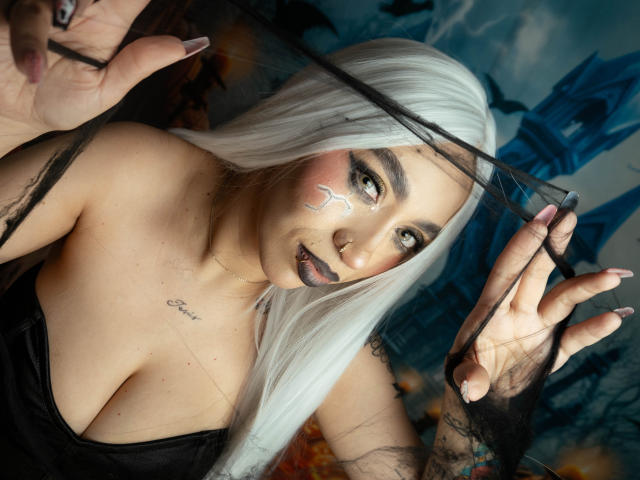 CamilleJuicyHot with long white hair, dark makeup, and a black top, holding black mesh, showing tits, tattoo on chest, and tattoos on hands, looking at camera.