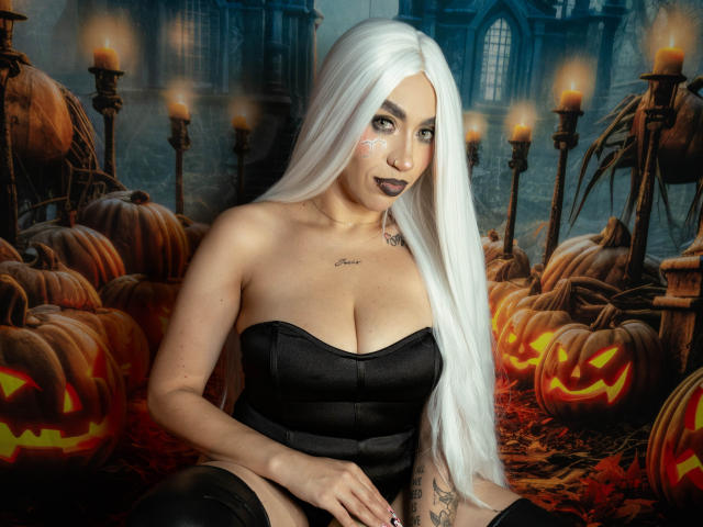 CamilleJuicyHot with long white hair, black strapless dress, dark lipstick, tattoos, and face paint, sitting among glowing pumpkins and candles, showcasing her tits and tattooed arm.