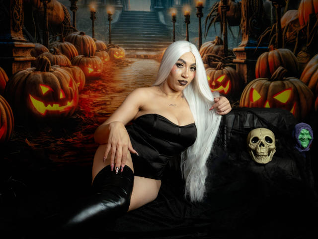 CamilleJuicyHot with long white hair, black strapless dress, and black boots, lying down with a pumpkin, skull, and green face on a black cloth. Her tits are prominent.