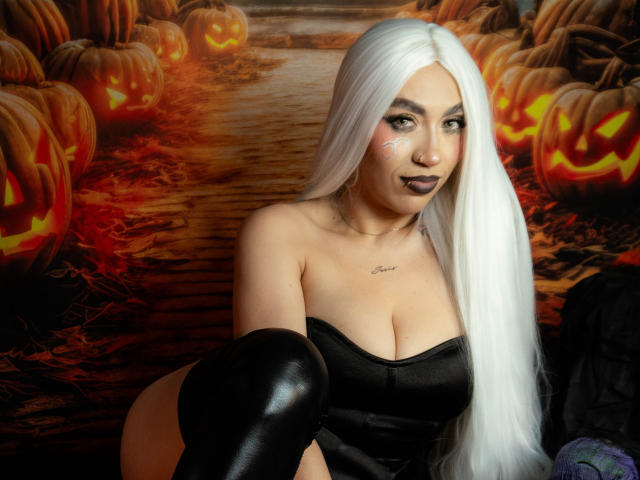 CamilleJuicyHot with long white hair, black lipstick, and white face paint, wearing a black strapless top and black thigh-highs, showing her big tits and a cute smile.