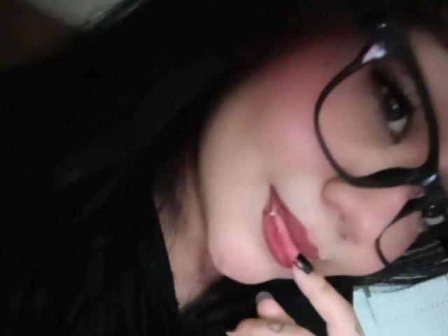 MalloryRamos with black glasses, pink blush, and glossy lips, touching her bottom lip with a finger, has long black hair, and a slightly mischievous smile.