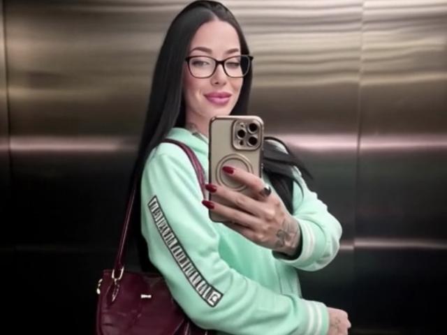MirandaHott69 with long black hair, glasses, mint green hoodie, red purse, taking selfie in elevator, holding gold phone, smiling, pink lipstick, tattoo on right hand.