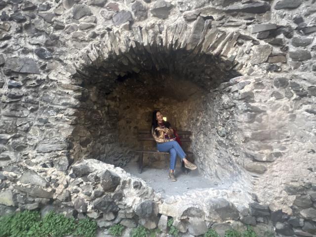 MirandaHott69 with small tits, sitting in a stone alcove, holding a cup, wearing blue jeans and brown boots, against a rough, gray stone wall.
