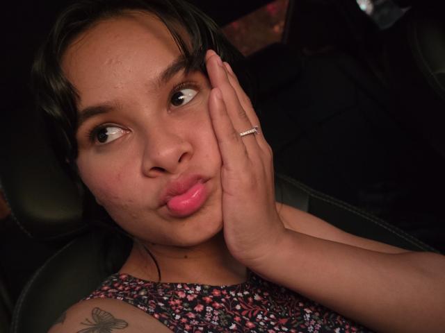 Yurimy with puffy pink lips, brown eyes looking sideways, hand covering part of her face, wearing a floral dress, ring on her finger, butterfly tattoo on her shoulder.