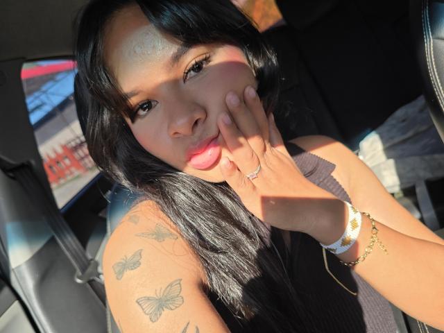 Yurimy with long black hair, pink lips, and tattoos on her arm, touches her cheek with a hand wearing a ring and a bracelet, sitting in a sunlit car.