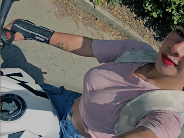 Yurimy with red lipstick, wearing a pink shirt and blue jeans, holding a motorcycle handle, wearing a black glove, visible tattoo on her arm, sunny day.