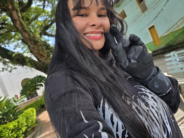 Yurimy with long black hair, smiling, wearing a black graphic shirt, black gloves, showing a finger to her lips, with visible tits under the shirt. Outside, green trees.