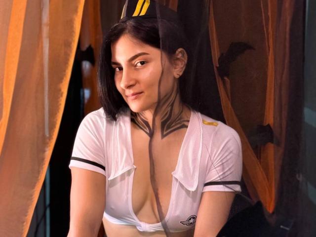LouissaBennet with black hair, wearing a white, sheer, low-cut, short-sleeve top, exposing her tits, and a bat tattoo on her neck, smiling subtly.