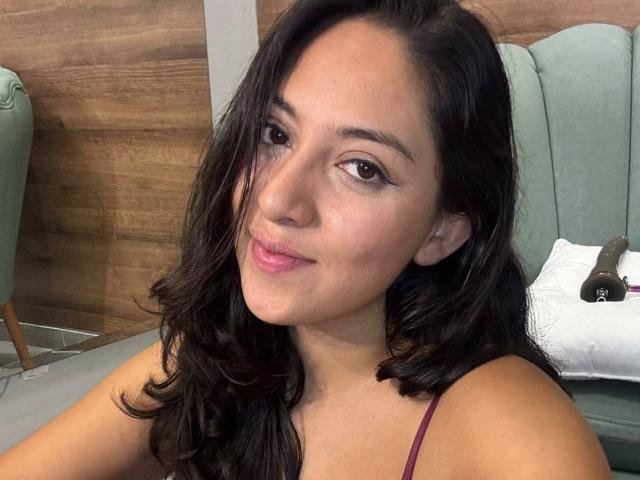 VictoriaLusty with wavy dark hair, brown eyes, pink lipstick, and a slight smile, wearing a maroon top, sitting in a green chair, looking at the camera.