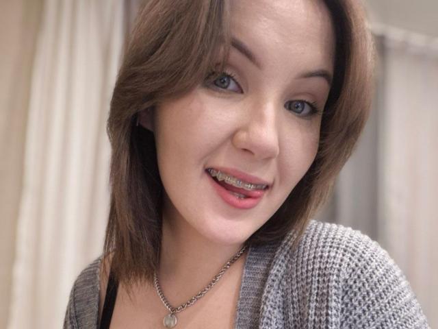 LidiaOckimey has brown hair, blue eyes, and a metal nose ring. She's smiling with metal braces, wearing a gray knit cardigan and a silver necklace.