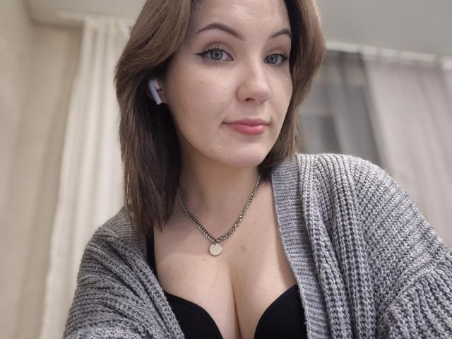 LidiaOckimey with medium brown hair, blue eyes, and pink lipstick, wearing a gray knitted cardigan over a black bra, showing her big tits. She has a necklace with a round pendant.