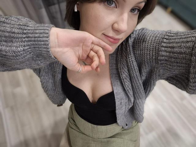 LidiaOckimey, with gray knitted cardigan, black bra showing her boobs, green high-waist pants, and hand near face, has fair skin, brown hair, and a slight smile.