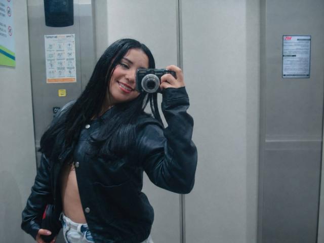 IsabellaCutte with long black hair, smiling, holding a camera, wearing an open black leather jacket, showing a bit of her flat stomach, standing in an elevator.