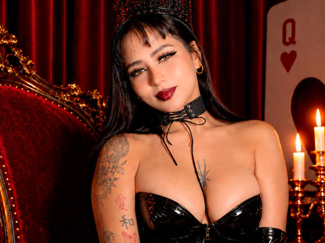Sophia, avec des seins en latex noir, des tatouages sur son bras droit, portant une couronne scintillante et un collier, du rouge à lèvres, et des boucles d'oreilles dorées, assise sur une chaise en velours rouge..
