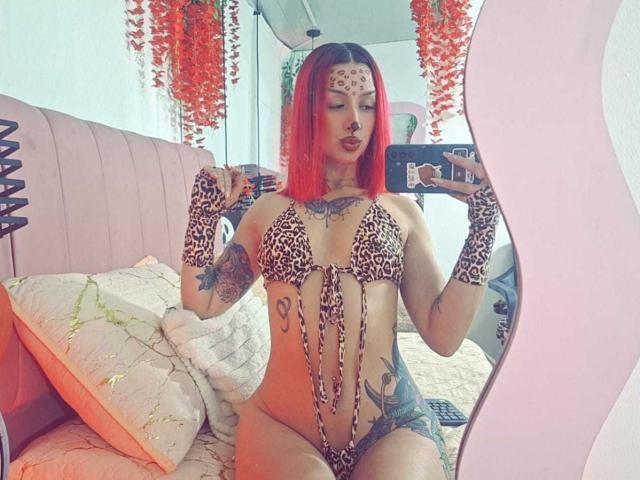 AnaRossii with red hair, leopard print bikini, fingerless gloves, tattoos, and face paint, taking a mirror selfie, showing her tits and pussy, sitting on a pink bed.