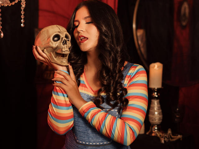 VelisseNoir with long black hair, holding a skull, tongue out, wearing a striped shirt and blue apron, candle in background, in a dark, moody setting, looks sexy.