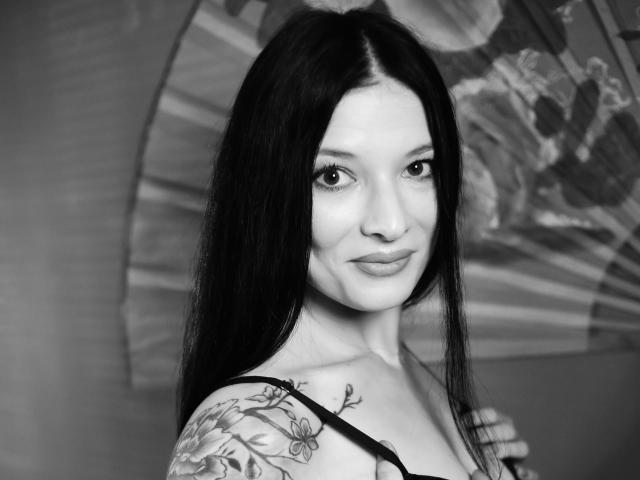 Black-and-white photo of MiaaCherry with long dark hair, light skin, tattoo on left shoulder, smiling, wearing a strap, subtle makeup, looking at camera.