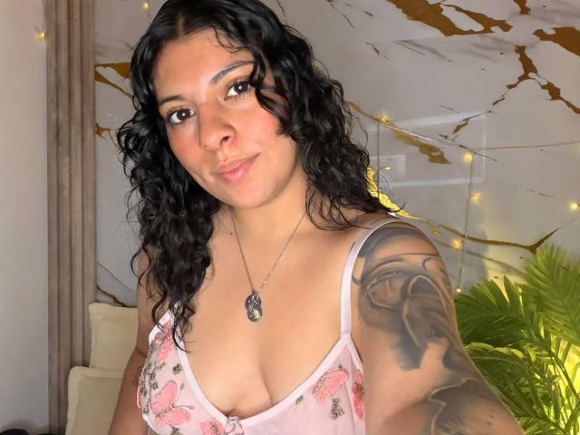 IsabellaFleur with curly black hair, light brown skin, and a tattoo on her right arm, wearing a pink floral top, showing her small tits and a subtle smile.
