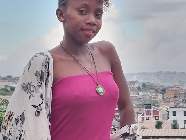 ShySexy, dark-skinned woman with small tits, wearing a pink strapless top and floral shawl, green pendant, standing outside, looking slightly to the side with a subtle smile.