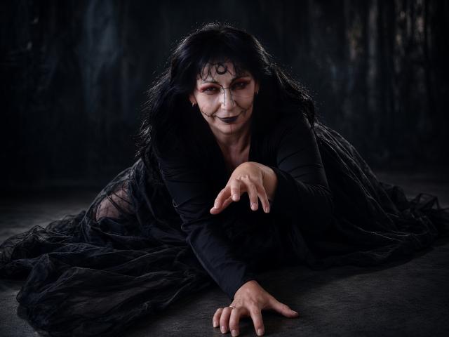 LiluGrants has black makeup, long hair, and a gothic outfit with a sheer black dress, crouching with a pointed finger and a sly grin. Dark, dramatic lighting.