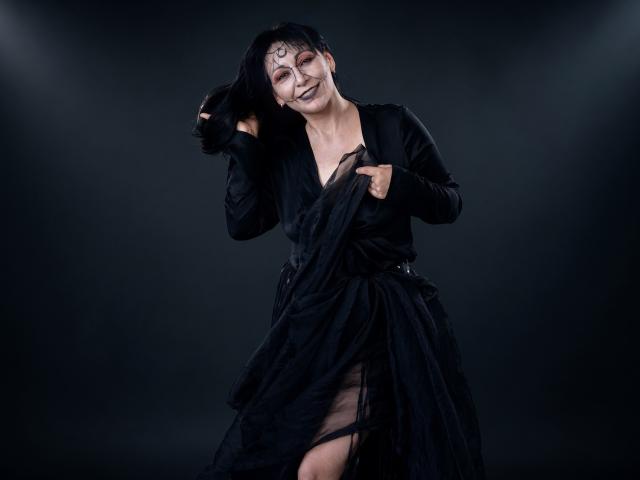 LiluGrants with black face paint, long black hair, and a black dress, holding her dress up to reveal her black panties, smiling, and looking slightly to the side.