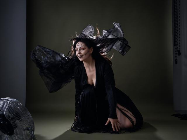LiluGrants kneels on the floor, black dress with deep cleavage, sheer black fabric flying around her head, dark makeup, next to a silver fan, looking sexy.