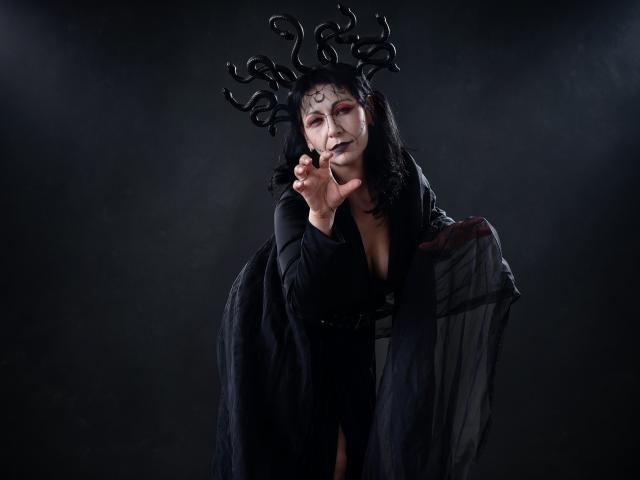 LiluGrants with dark makeup, black hair, and antler headpiece, wearing a black robe, holding her chin, and draped in a sheer cloth over her tits. Dark, moody photograph.