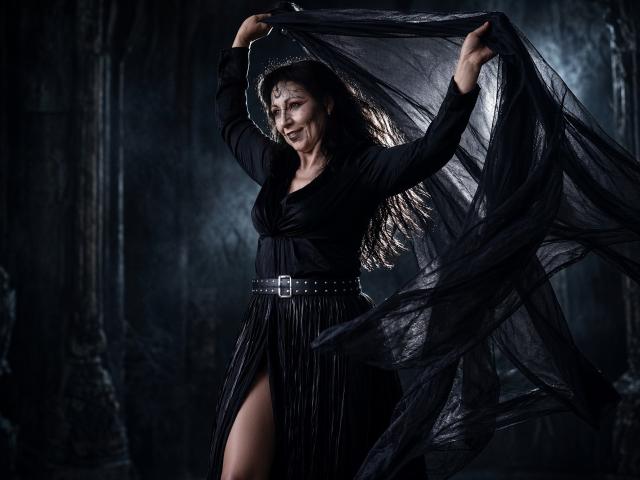 LiluGrants with black dress, holding sheer black fabric, smiling, arms raised, belt with buckle, thigh slit, dark wavy hair, dim lighting, looking sexy, black background.