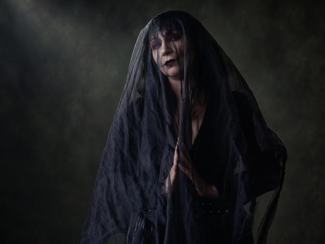 LiluGrants with dark makeup, wearing a black sheer veil, exposing her tits, holding the veil, looking sinister, in a moody, low-light photograph.