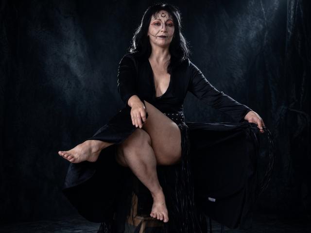 LiluGrants with dark hair, black dress, and face paint, sitting with one leg up, exposing her bare foot and thigh, her tits visible, black background, looking at camera.