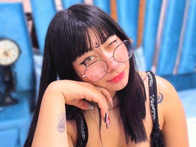Genesiss with black hair, glasses, and tattoos, winking, resting her chin on her hand, wearing a black bra, showing her tits, in a blue room.