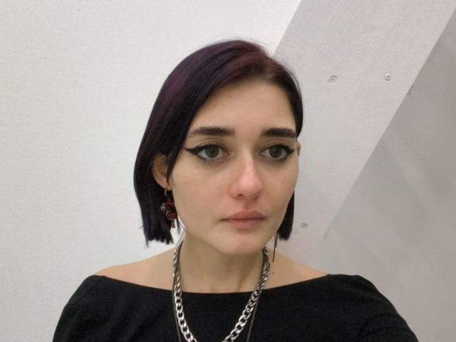 Ligia Gottshall, avec ses courts cheveux violets, son haut noir à une épaule, son eye-liner épais, son collier en chaîne et ses boucles d'oreilles rouges, regarde droit devant la caméra avec une expression neutre..