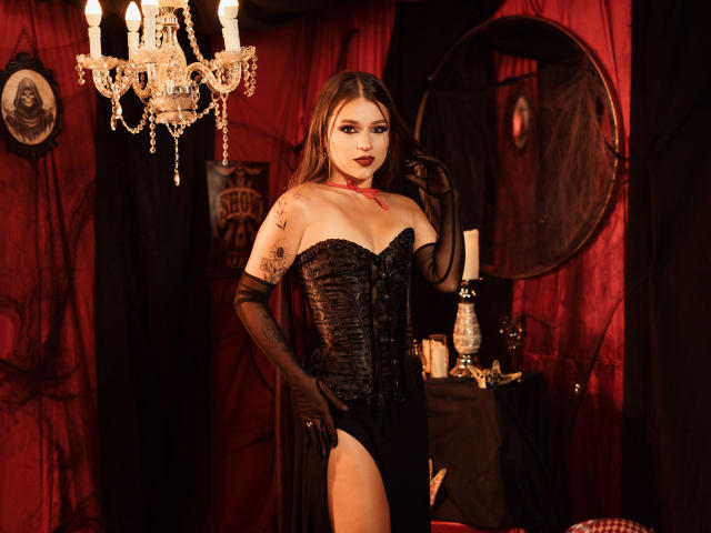 MilanaEvan with long brown hair, wearing a black lace corset, black gloves, and a red choker, standing in a dimly lit room with red curtains and a crystal chandelier.