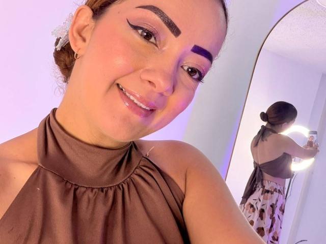 EimmyLove with brown halter top, thick black eyebrows, purple eyeliner, and shiny lips, smiling in mirror selfie, showing back in floral pants, holding phone with ring light.