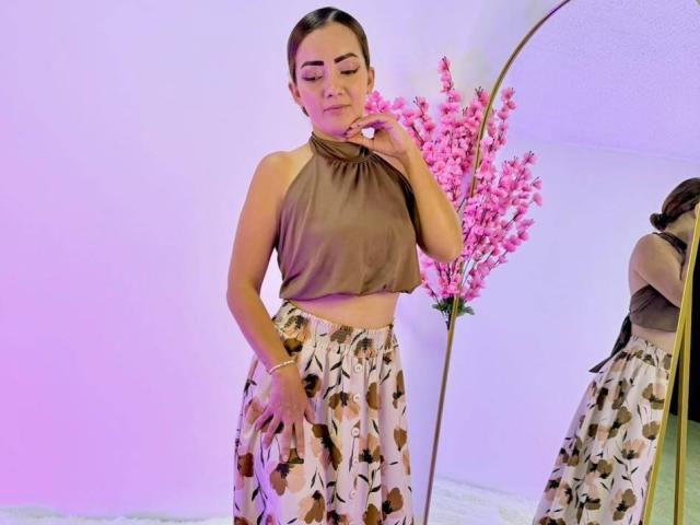 EimmyLove with brown halter top, floral skirt, and pink flowers in mirror, touching chin with hand, reflecting in mirror, wearing bracelet, standing in softly lit room.